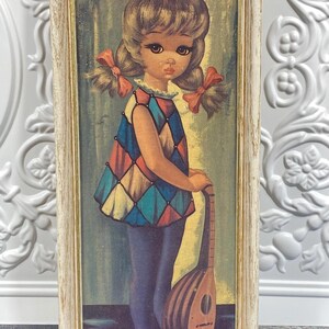 May include: A framed wall art print featuring a young person with pigtails and red bows, wearing a sleeveless harlequin-patterned tunic in blue, red, and yellow, and dark blue pants. The figure holds a wooden lute-style stringed instrument.