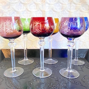 May include: A collection of six colorful wine glasses with etched starburst designs. The glasses feature a variety of colors, including red, purple, green, yellow, and blue. Each glass has a clear stem and base, and they are arranged on a black tray.