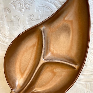 Puede incluir: Una fuente de servir vintage en forma de hoja con tres compartimentos. La fuente tiene un esmalte marrón brillante con reflejos más claros. El diseño recuerda a la vajilla moderna de mediados de siglo.