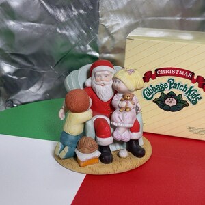 May include: A Cabbage Patch Kids Christmas figurine featuring Santa Claus sitting in a light green armchair with a child kneeling beside him and another child sitting on his lap holding a small teddy bear, accompanied by a yellow product box.