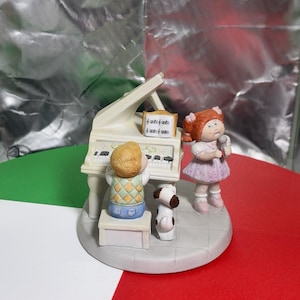 May include: A ceramic figurine set featuring a child playing a white grand piano, a child with red hair standing and holding a microphone, and a small dog. The piano has a music sheet stand with printed notes. The figures are on a circular base.