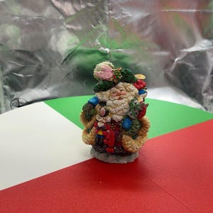 May include: A decorative Santa Claus figurine featuring a white beard, a green hat adorned with a pink feather and red berries, and a tan fur-trimmed coat. The figure holds a small nutcracker and a sack of gifts, standing on a textured base.
