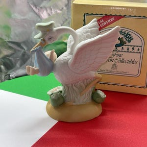 May include: A white porcelain stork figurine wearing a light green cap and carrying a light blue bundle with a baby inside. The stork stands on a tan base with green foliage and white flowers. A yellow box with the text Patch Kids Fine Collectibles.