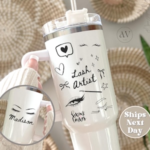 May include: White tumbler with a handle, decorated with black eyelash and heart illustrations, and the words "Lash Artist." A smaller tumbler shows the name "Madison" with eyelashes. The tumbler has a white lid and a straw.