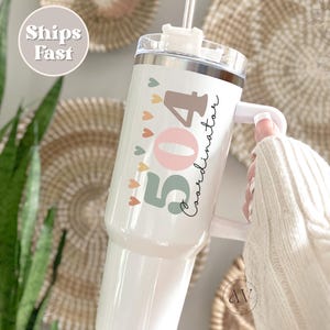May include: White stainless steel tumbler with a handle and a straw. The tumbler has a design with the numbers 504 and the word "Coordinator" written in cursive. The numbers are in different colors, including pink, green, and brown. There are small hearts around the numbers.