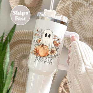 May include: White tumbler with a Halloween-themed design. The design features a ghost, pumpkin, and floral accents. The text "Mrs. Willis" is written on the tumbler. The tumbler has a handle and a straw.