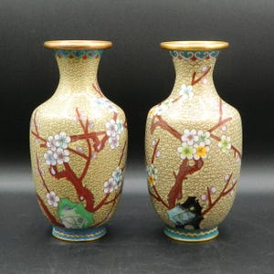 Pode incluir: Um par de vasos de cloisonné amarelo com um design floral. Os vasos são decorados com um padrão de flores brancas e rosas sobre um fundo amarelo. Os vasos medem aproximadamente 15 cm de altura.