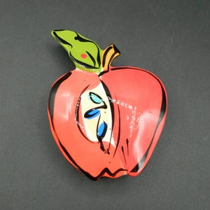 Puede incluir: Un plato de cerámica con forma de manzana roja con una hoja verde y un contorno negro. El interior de la manzana está pintado con detalles blancos, azules y verdes.