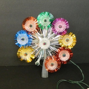 May include: A vintage Christmas tree topper featuring a silver starburst centre and colourful, metallic flower-like accents in shades of blue, green, purple, yellow, orange, and red. The topper has a clear plastic base and a green electrical cord.