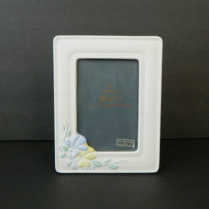 May include: A white ceramic picture frame with a rectangular aperture. The frame is decorated with a floral motif in blue, yellow, and green. The frame has a dark grey interior and a "Bone China" label.