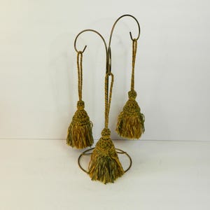 Könnte beinhalten: Ein dekorativer Metallständer mit drei hängenden Quasten. Die Quasten sind eine Mischung aus Grün und Gold, und der Ständer hat ein geschwungenes Design. Der Ständer hat einen kreisförmigen Sockel und einen Haken zum Aufhängen.