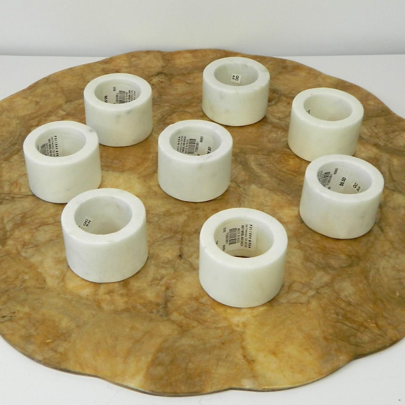 Puede incluir: Ocho servilleteros de m&aacute;rmol blanco dispuestos sobre una superficie decorativa y texturizada de color marr&oacute;n. Cada anillo es cil&iacute;ndrico con un acabado liso y pulido. Los servilleteros son de diferentes alturas y anchos.
