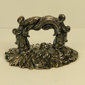 May include: A silver-toned metal drawer pull featuring an ornate, raised relief design of scrolling leaves and botanical motifs. The handle is arched with a textured surface, mounted on an oval-shaped base plate with matching intricate detailing.
