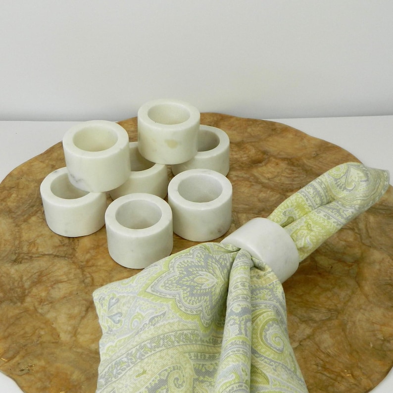 Puede incluir: Un conjunto de servilleteros de m&aacute;rmol blanco, de forma cil&iacute;ndrica, dispuestos sobre una superficie de madera. Un servilletero sujeta una servilleta plegada con un estampado verde y gris. Los anillos de m&aacute;rmol tienen un acabado liso y pulido.