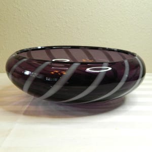 May include: A decorative glass bowl with a deep purple hue, featuring a spiral design of alternating purple and white stripes. The bowl has a rounded shape and a glossy finish, suitable for home decor.