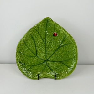 Op de afbeelding: Een groene keramische blad-vormige plaat met een getextureerd oppervlak en een klein rood lieveheersbeestje accent. De plaat heeft een gedetailleerd aderpatroon en wordt gepresenteerd op een wit oppervlak.