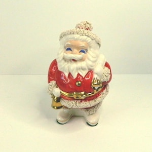 May include: A ceramic Santa Claus figurine featuring a glossy red coat with a gold belt, white textured trim, and a matching hat. The figure has a white beard, blue eyes, and holds a gold-colored bell in one hand while carrying a sack over one shoulder.