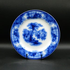 May include: A blue and white porcelain dinner plate with a scenic design. The plate features a blue and white landscape with a tree, a building, and a river. The plate has a blue and white floral border.