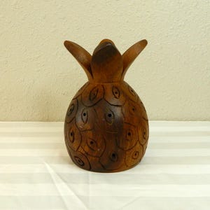 Könnte beinhalten: Eine geschnitzte Ananas-Figur aus Holz. Die braune Ananas hat eine strukturierte Oberfläche mit geschnitzten Details und einer blattartigen Krone. Die Figur wird auf einer weiß-grau gestreiften Oberfläche präsentiert.