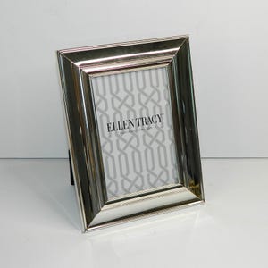 May include: A rectangular silver-toned metal picture frame with a beveled edge. The frame contains a display insert featuring a grey and white geometric pattern and the text ELLEN TRACY 4 inch x 6 inch / 10 cm x 15 cm centered on the display.