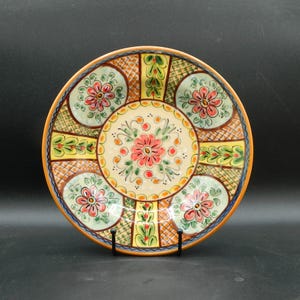 Op de afbeelding: Een rond keramiek bord met een kleurrijk bloemenontwerp. Het bord heeft een witte achtergrond met een centrale cirkel met een roze bloem met groene bladeren. De buitenrand van het bord is versierd met een herhalend patroon van gele, groene en oranje vierkanten en cirkels.