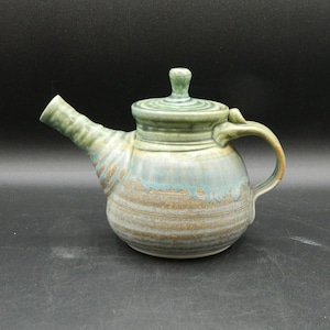 Pode incluir: Uma chaleira de cerâmica feita à mão com um esmalte verde e listras marrons. A chaleira tem um corpo arredondado, um bico curvo e uma tampa com uma pega.