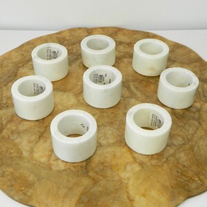 Puede incluir: Ocho servilleteros de m&aacute;rmol blanco dispuestos sobre una superficie texturizada de color marr&oacute;n claro. Cada anillo es cil&iacute;ndrico con una abertura circular. Algunos anillos tienen una peque&ntilde;a etiqueta. El m&aacute;rmol tiene un acabado liso y pulido.