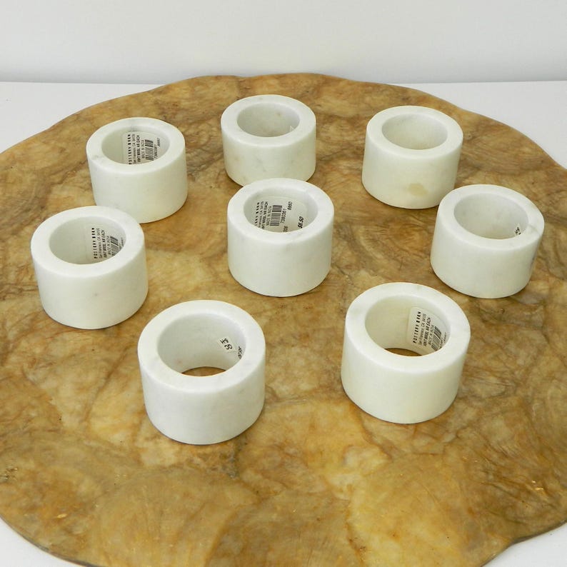 Puede incluir: Ocho servilleteros de m&aacute;rmol blanco dispuestos sobre una superficie texturizada de color marr&oacute;n claro. Cada anillo es cil&iacute;ndrico con una abertura circular. Algunos anillos tienen una peque&ntilde;a etiqueta. El m&aacute;rmol tiene un acabado liso y pulido.