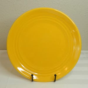 Puede incluir: Un plato de cerámica amarillo brillante con detalles de anillos concéntricos. El plato es redondo y tiene un acabado brillante. El plato se exhibe en un soporte de plato de metal negro. Este plato es adecuado para servir alimentos.