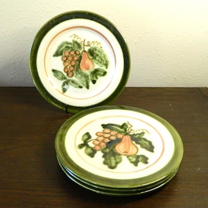 May include: A set of ceramic plates with a hand-painted fruit design. The plates feature a white center with a brown border and a green rim. The fruit design includes grapes and a pear. The plates are stacked on a dark wooden surface.
