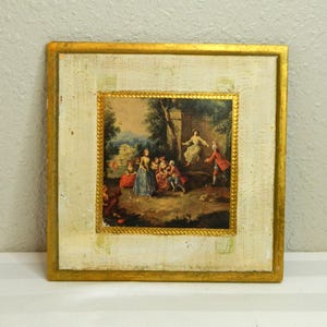 May include: Square framed artwork featuring a classical painting of people in a garden setting. The painting is framed with a gold border and set within a larger off-white frame with a gold trim. The artwork depicts figures in period clothing.