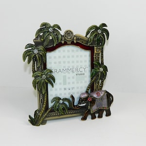 May include: A rectangular metal picture frame featuring a dark bronze finish with sculpted palm trees adorned with clear crystals. A decorative elephant with a purple and gold blanket stands at the base. The insert reads Grammercy Studio 3.5" x 5".