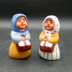 May include: Two ceramic figurines of women, each wearing a headscarf and holding a basket. One figurine has a blue headscarf with red polka dots, the other has a white headscarf with red polka dots. Both have yellow and orange clothing.