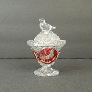 May include: A clear glass candy dish with a lid and a bird-shaped handle. The dish has a red and clear design with a bird motif. The dish is likely vintage or antique.