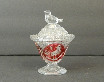May include: A clear glass candy dish with a lid and a bird-shaped handle. The dish has a red and clear design with a bird motif. The dish is likely vintage or antique.