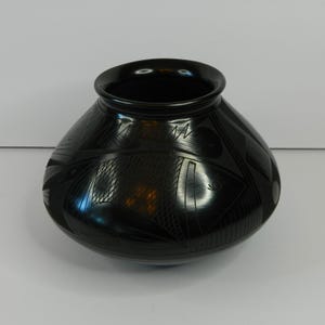 May include: A black ceramic pot with a rounded body and a slightly flared rim. The pot features intricate geometric designs, including triangles, circles, and lines, etched into the surface. The glossy finish enhances the dark color and the detailed patterns.