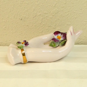 May include: A light pink porcelain hand-shaped dish with a gold band around the wrist. The dish is decorated with purple and yellow pansy flowers and green leaves. A decorative item.