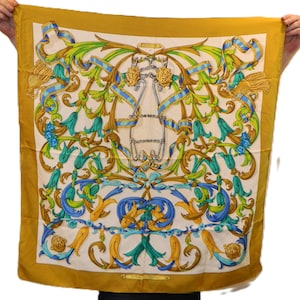 May include: A square silk scarf featuring a mustard yellow border and a central cream panel. The design displays intricate blue and green scrolling acanthus leaves, golden tassels, and a central silver horse bit motif with blue ribbons.