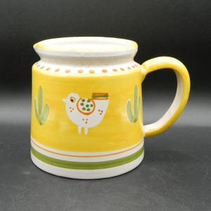 Könnte beinhalten: Gelbe Keramiktasse mit weißem Rand und Griff. Die Tasse hat ein handgemaltes Design eines weißen Vogels mit orangefarbenen und grünen Akzenten sowie grüne Kaktusillustrationen. Der Boden hat grüne und orangefarbene Streifen.