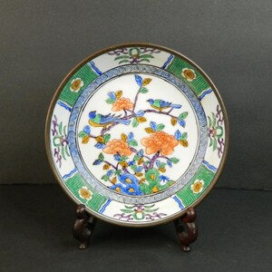 May include: A round ceramic plate featuring a central design of two blue and yellow birds perched on branches with orange flowers and green leaves. The border has a green striped pattern with small white flowers and blue wavy lines.