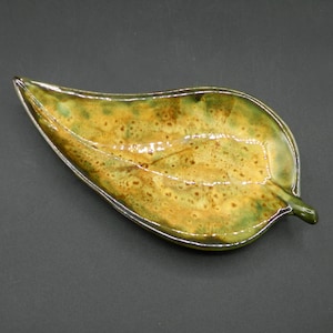 May include: A ceramic leaf-shaped dish with a green and yellow glaze. The glaze has a speckled appearance, with brown spots throughout.