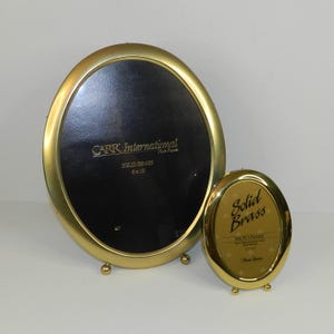May include: Two oval-shaped photo frames made of solid brass with a polished gold finish. The larger frame features text reading CARR International SOLID BRASS 8x10, while the smaller frame displays the text Solid Brass PHOTO FRAME 5x7.