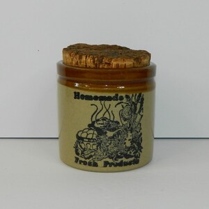 Può includere: Un barattolo di ceramica beige con bordo marrone e coperchio in sughero. Il barattolo presenta un'illustrazione nera e le parole "Homemade Fresh Products". Il barattolo è cilindrico e poggia su una superficie bianca.