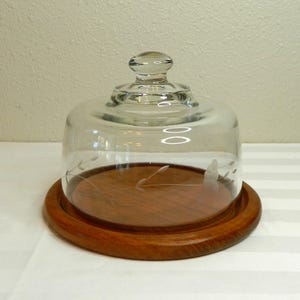 Puede incluir: Una campana de cristal transparente con una base de madera redonda. La cúpula de cristal tiene un pequeño asa en la parte superior y diseños florales grabados. La base de madera es de color marrón oscuro, y la campana está diseñada para cubrir alimentos.