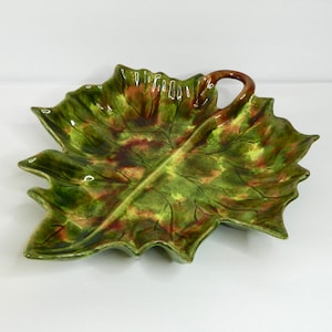 May include: A ceramic leaf-shaped serving dish with a handle. The dish features a glossy finish with shades of green, brown, and yellow. The leaf design includes detailed veining and a curved handle. This decorative piece is suitable for serving snacks or as a decorative accent.