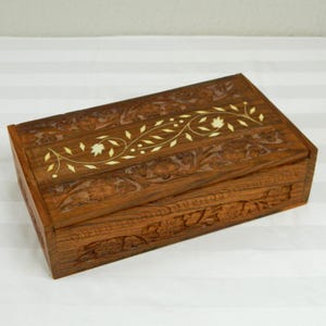 May include: A rectangular wooden box with a hinged lid. The box is a warm brown color and features intricate floral carvings. A decorative ivory-colored inlay of leaves and flowers adorns the lid, adding a touch of elegance. The box is likely used for storing small items.