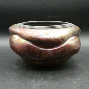 Könnte beinhalten: Eine handgefertigte Keramikschale mit einer einzigartigen, strukturierten Oberfläche. Die Schale hat eine reichhaltige Farbpalette aus Burgunder, Bronze und Gold mit einem dunklen Rand. Die abgerundete Form und das schillernde Finish verleihen ihr ein unverwechselbares, künstlerisches Aussehen.