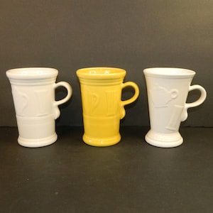 May include: Three ceramic mugs arranged in a row, featuring two white mugs on the ends and one yellow mug in the centre. Each mug has a flared rim, a curved handle, and a raised embossed design on the exterior surface depicting a teapot and a spoon.