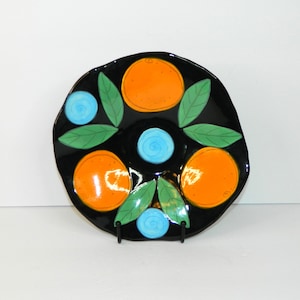 May include: A black ceramic plate with a vibrant design featuring orange circles, green leaves, and blue spiral accents. The plate has a glossy finish and is supported by a black metal stand. The design is reminiscent of citrus fruit.