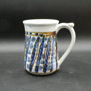 Może przedstawiać: Biały kubek ceramiczny z abstrakcyjnym wzorem w kolorze niebieskim, brązowym i białym. Kubek ma zaokrąglony korpus i uchwyt.